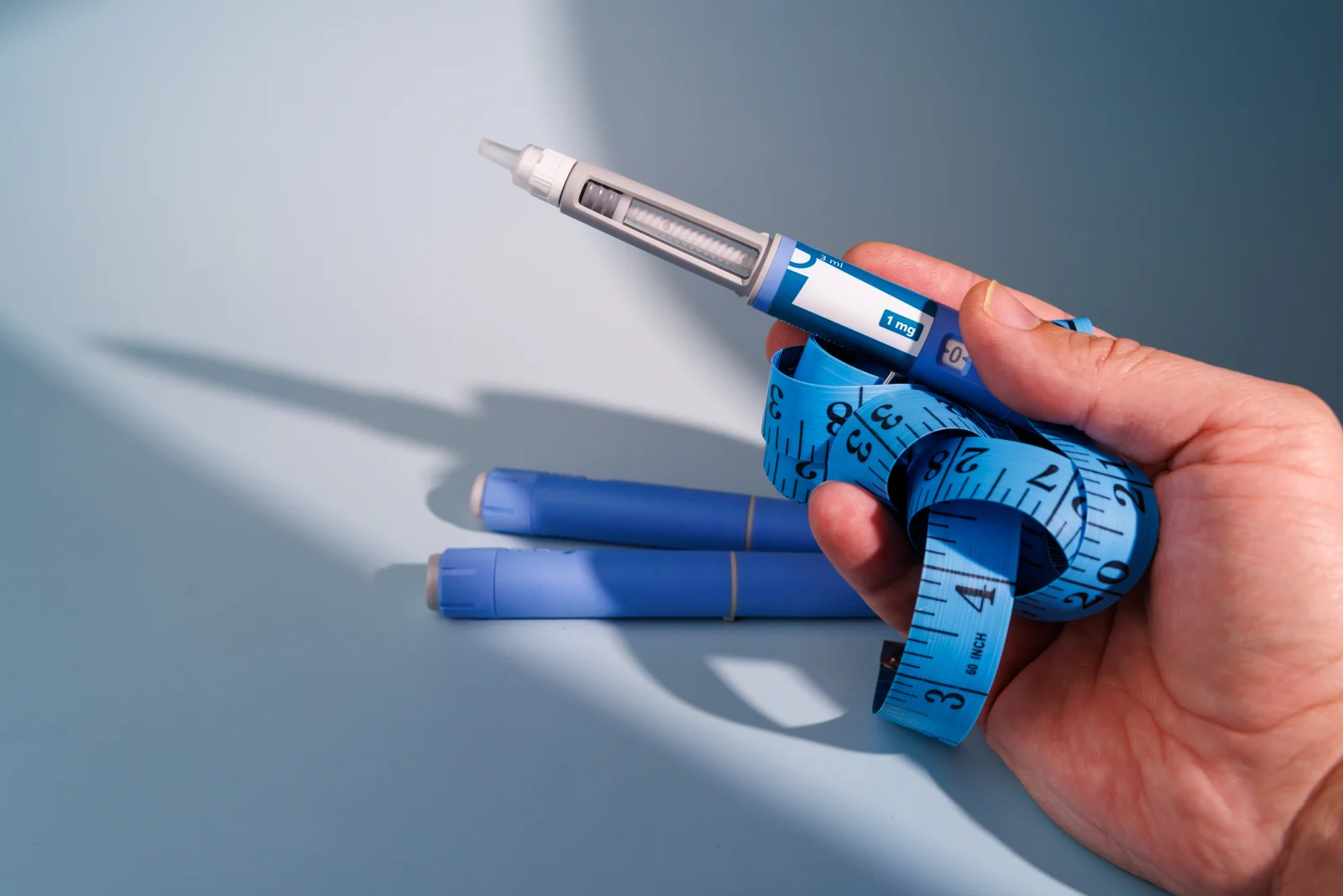 Hand holding a 1 mg semaglutide injection pen with blue measuring tape and two additional pen cartridges